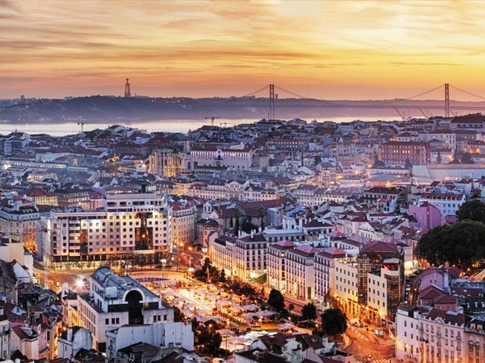 Vista panorámica de Lisboa con tejados rojos, el río Tajo y el puente 25 de Abril al fondo, bajo un cielo despejado