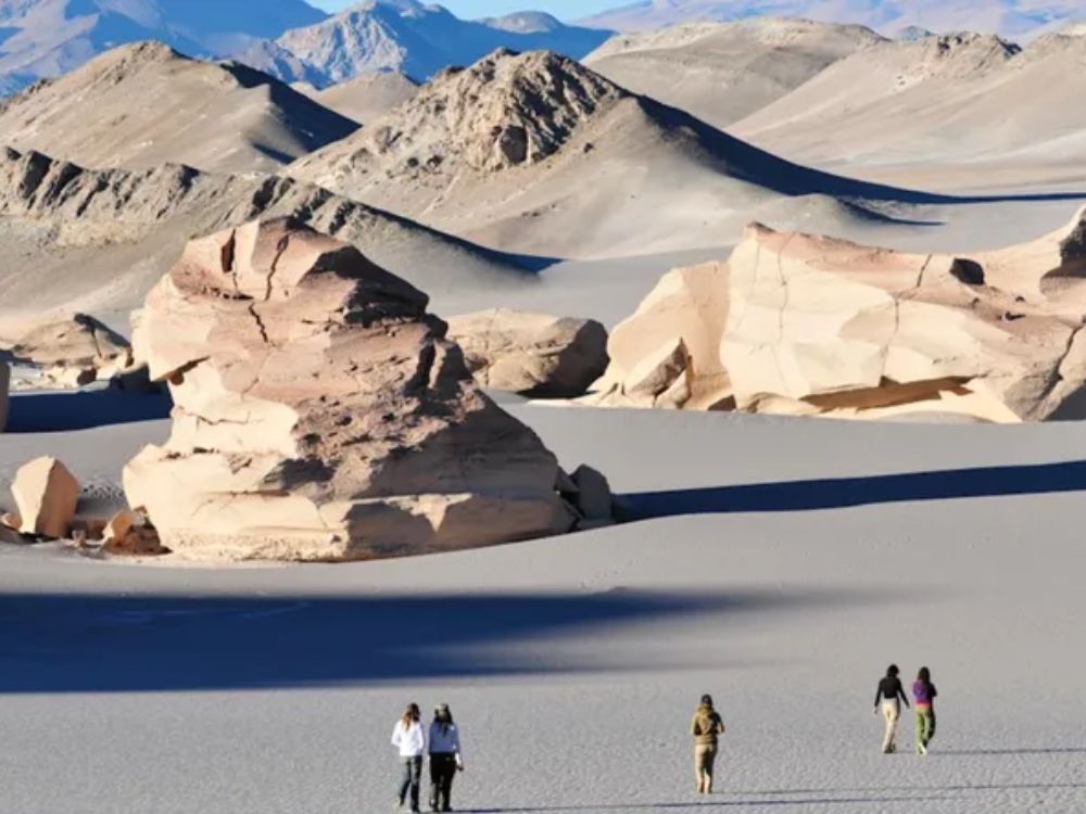 Grupo de personas caminando entre formaciones rocosas blancas en el paisaje desértico de Campo de Piedra Pómez, con montañas al fondo