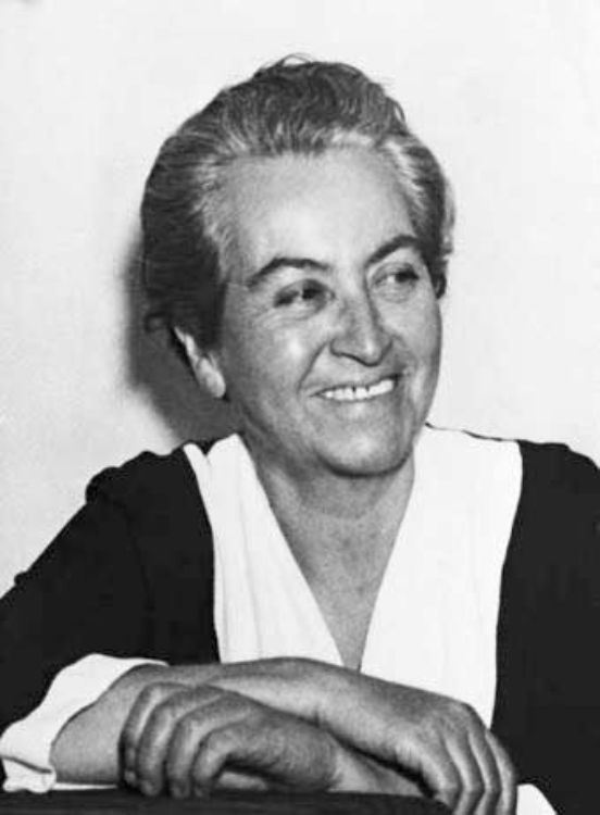 Retrato en blanco y negro de Gabriela Mistral, sonriente, con el cabello corto peinado hacia atrás y los brazos apoyados sobre una superficie
