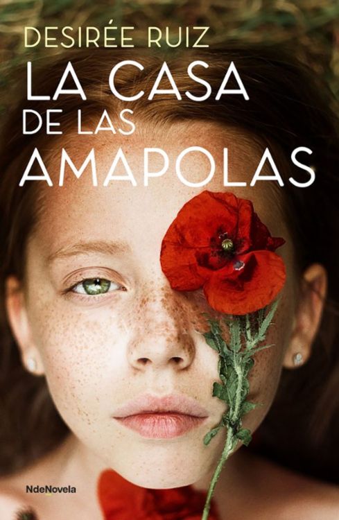 'La casa de las amapolas'. Rostro joven con piel clara y pecas, tendido sobre hierba; una amapola roja cubre uno de sus ojos, evocando misterio y belleza