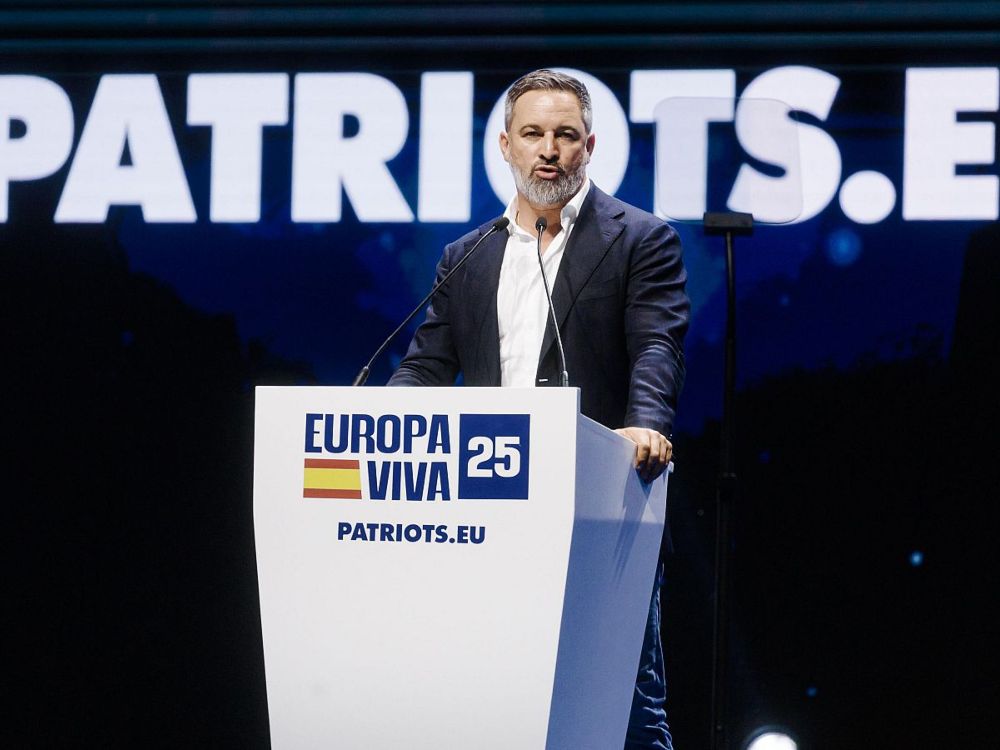La etiqueta como arma. Santiago Abascal en el escenario de Europa Viva 25, hablando desde un atril con el lema PATRIOTS.EU proyectado al fondo
