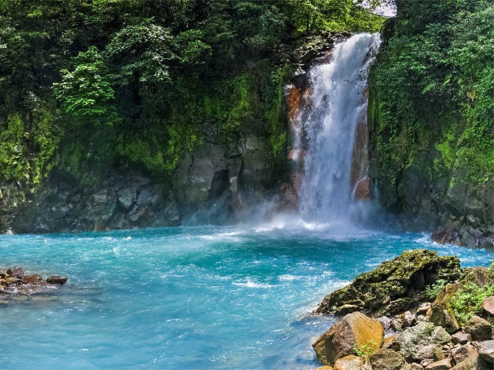 Cascada del Río Celeste cayendo sobre una poza turquesa, rodeada de vegetación tropical y rocas húmedas