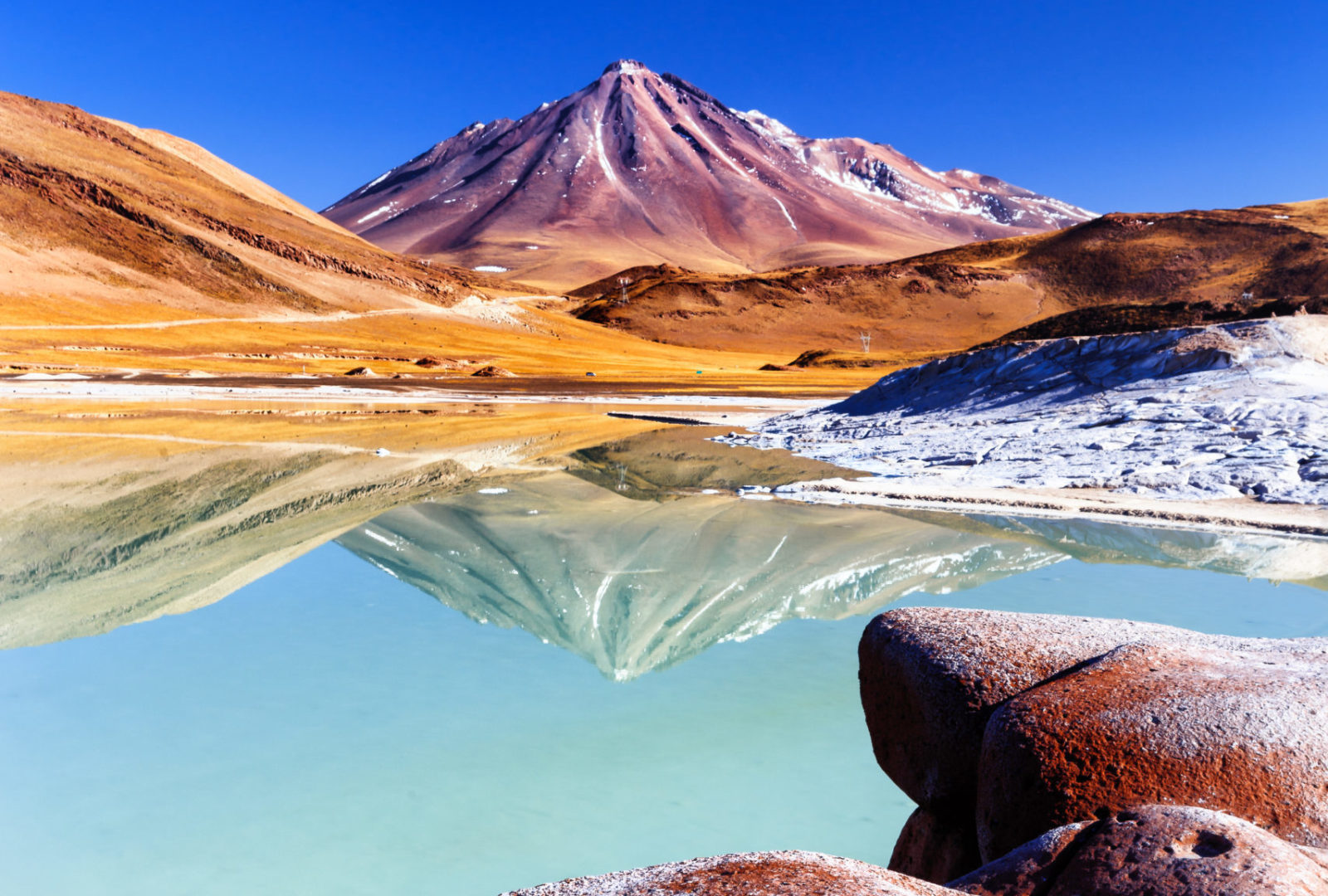 Salar de Tara. Paisaje altiplánico con volcán nevado reflejado en laguna turquesa, rodeado de depósitos minerales y terreno árido