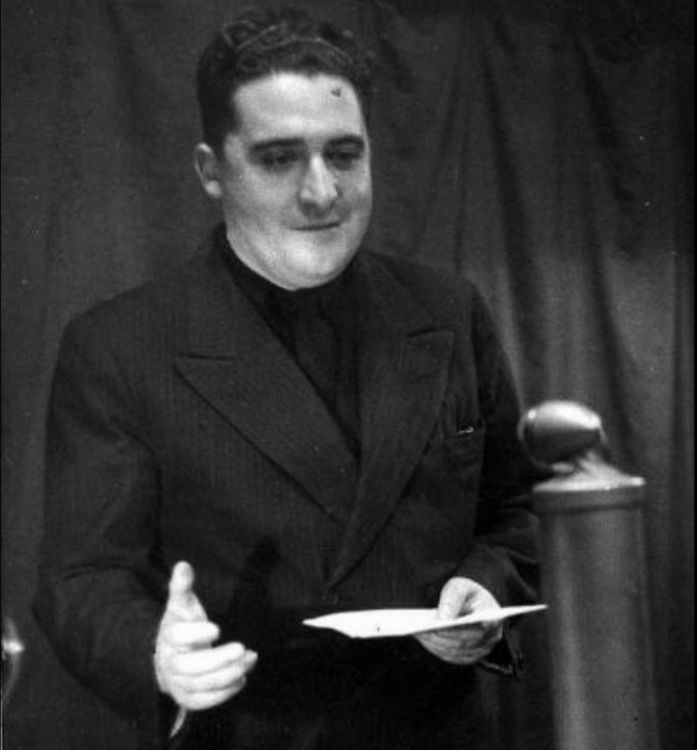 Agustín de Foxá RAE Agustín de Foxá, de pie en un acto oficial, vestido con traje oscuro, sosteniendo papeles y gesticulando