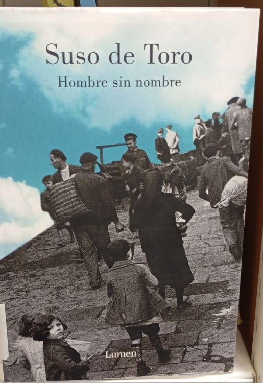 Cubierta de 'Hombre sin nombre', de Suso de Toro. Fotografía de varias personas ascendiendo por una calle empedrada, con cielo en cian brillante.