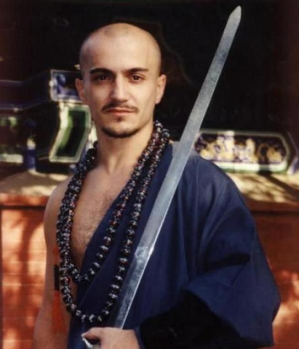 Aguilar, el falso monje, con la cabeza afeitada sosteniendo una espada vertical frente a un fondo arquitectónico de estilo oriental