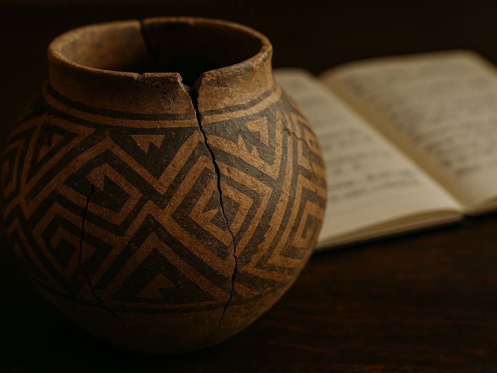 Vasija arikara rota con motivos geométricos sobre mesa oscura, junto a cuaderno desenfocado con anotaciones lingüísticas