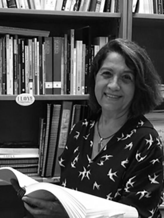 Las siete virtudes capitales. Márgara Russotto de pie frente a una estantería llena de libros, sosteniendo un libro abierto y mirando a cámara