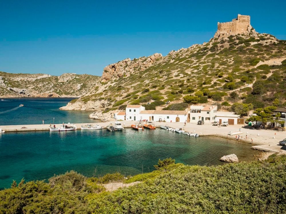 Vista del puerto de Cabrera con aguas turquesas, casas blancas y el castillo en lo alto del promontorio