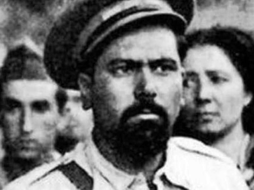 Retrato en blanco y negro de Valentín González 'El Campesino' con uniforme militar, rodeado de varias personas en segundo plano