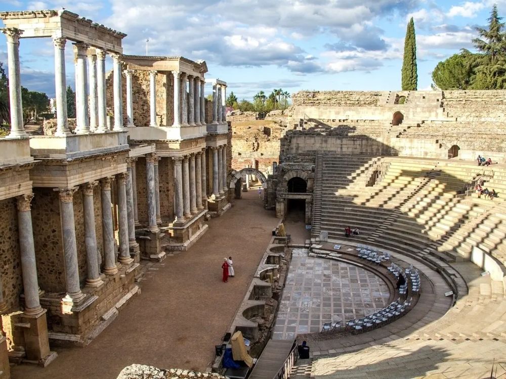 Emeritenses Emeritenses. Teatro Romano de Mérida con su graderío semicircular y la escena monumental de columnas corintias, con varios visitantes recorriendo el conjunto arqueológico