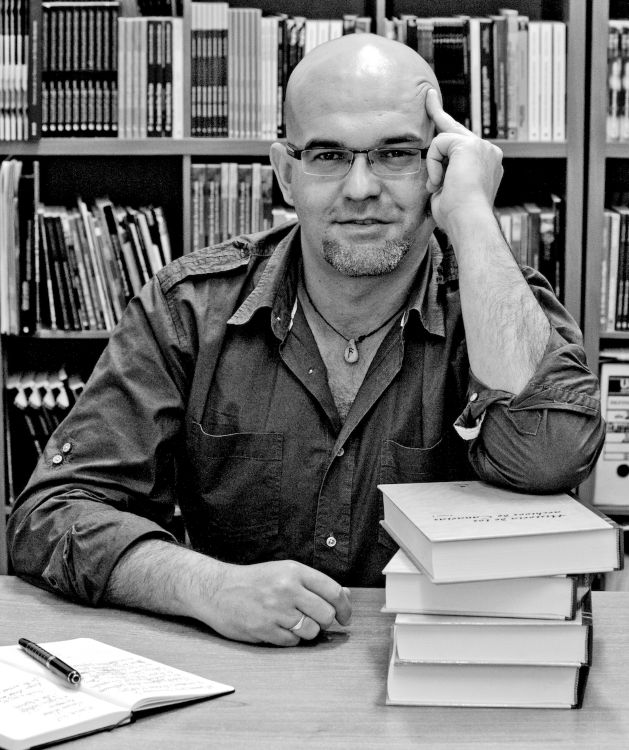'Filologías', Retrato en blanco y negro de Alexis Ravelo sentado ante una mesa con libros y un cuaderno, en una biblioteca