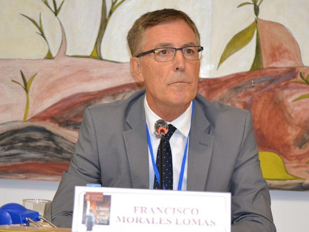'Bendita la eternidad'. Francisco Morales Lomas, sentado ante un micrófono en una mesa de conferencia, con un mural colorido al fondo