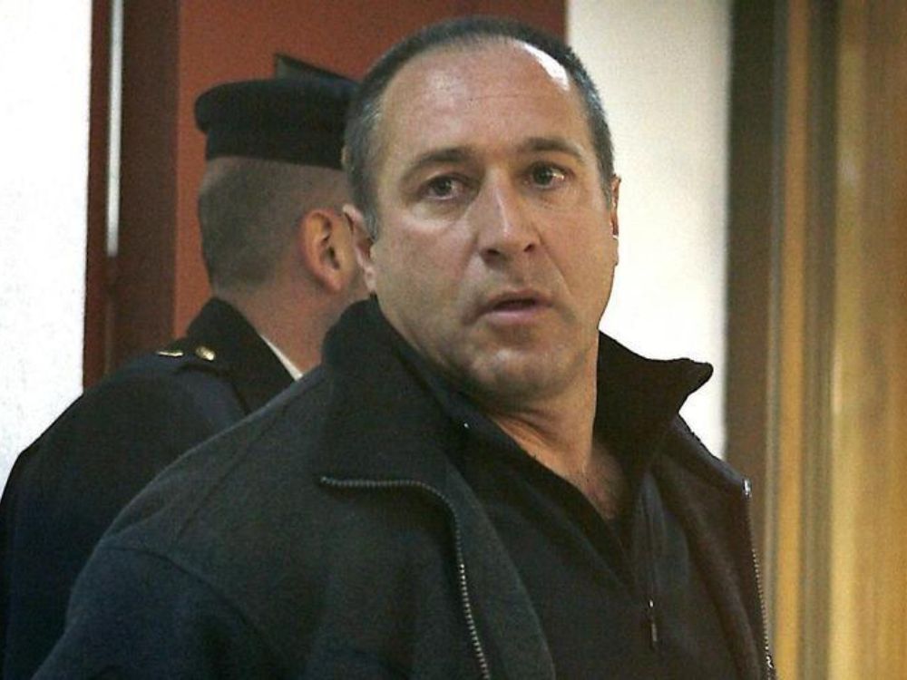 Retrato de Henri Parot escoltado por un agente en un pasillo judicial, mirando a cámara con expresión desafiante