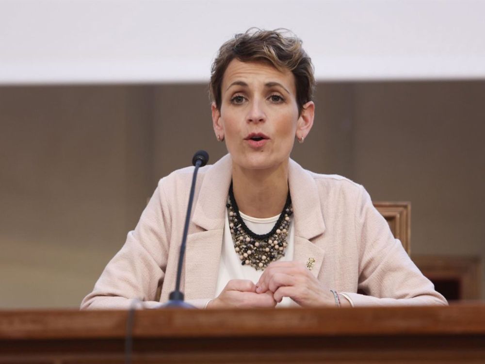 María Chivite hablando al micrófono, con chaqueta clara, collar de cuentas y un vaso de agua a su lado