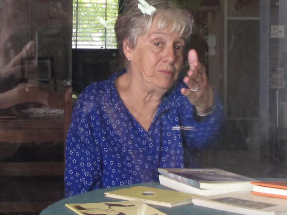 Circe Maia sentada ante una mesa con libros abiertos, gesticulando mientras conversa, vista a través de un cristal que refleja parcialmente la escena.