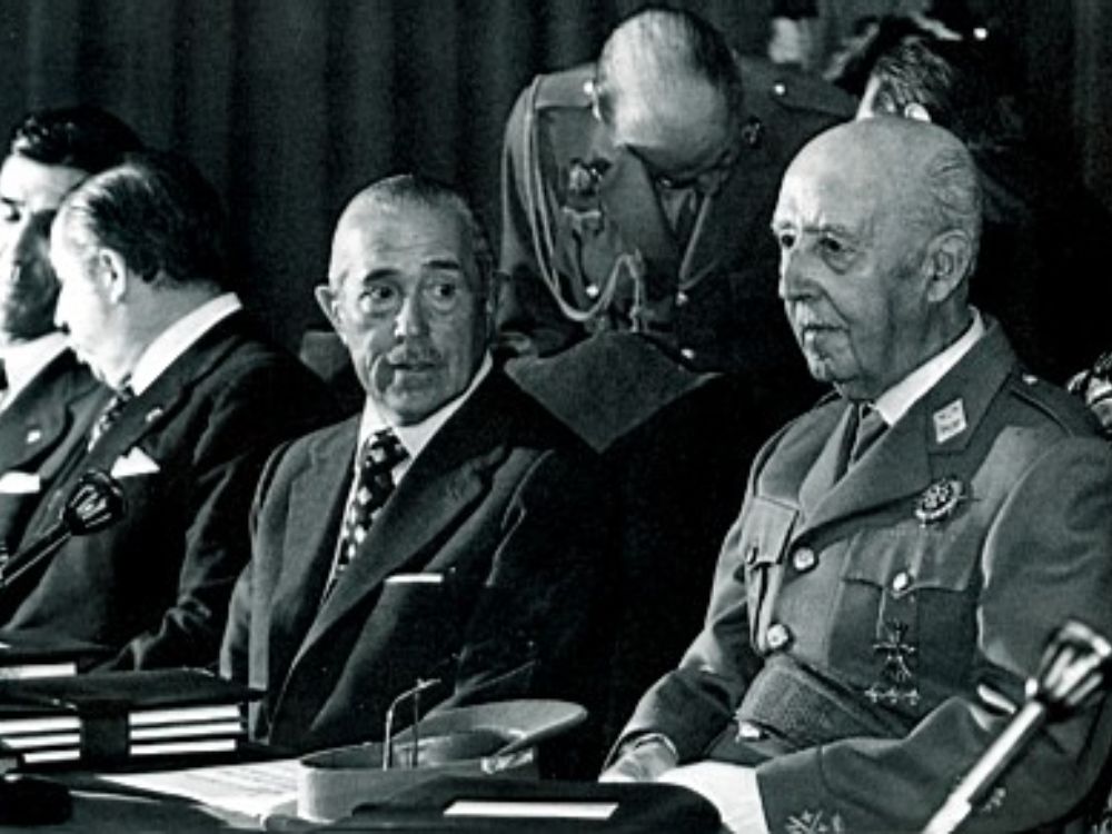 El espíritu del 12 de febrero. Fotografía en blanco y negro de Francisco Franco y Carlos Arias Navarro sentados en una mesa durante un acto oficial