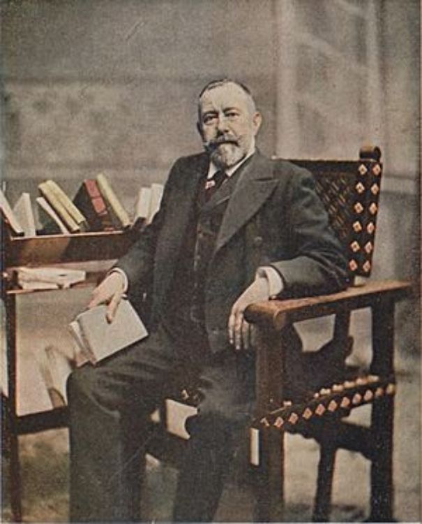 Retrato de Eugenio Sellés, sentado en un sillón, con barba y traje oscuro, sosteniendo un libro abierto junto a una mesa con varios volúmenes