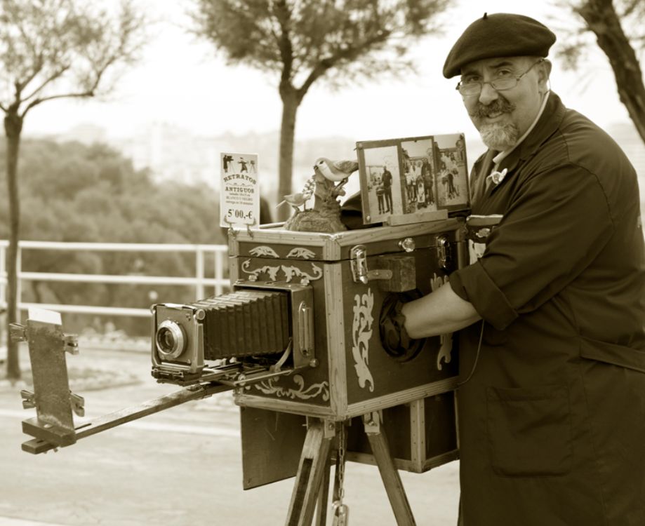 Fotógrafo minutero sonriente operando una cámara de cajón decorada en una plaza, con muestras de retratos antiguos y cartel de precios