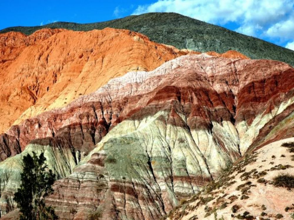 Quebrada de Humahuaca Vista del Cerro de los Siete Colores en Purmamarca, dentro de la Quebrada de Humahuaca, con montañas de tonos rojos, verdes, rosados y ocres bajo un cielo parcialmente nublado