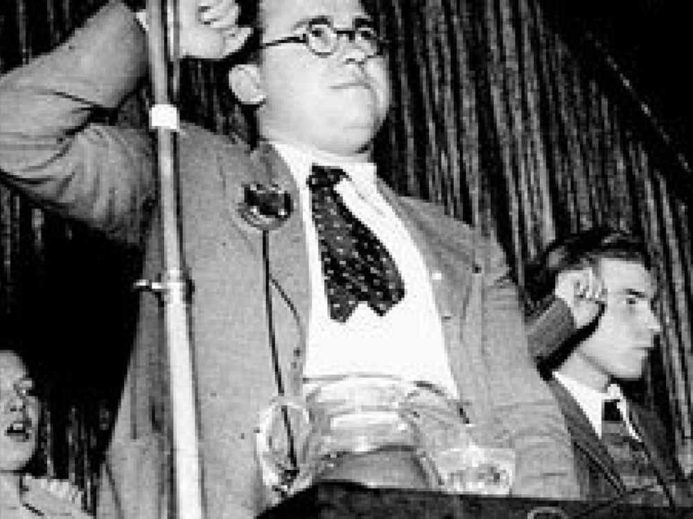 Santiago Carrillo, joven y con gafas, de pie a la izquierda junto a otros militantes, todos con el puño en alto frente a un micrófono en un acto político